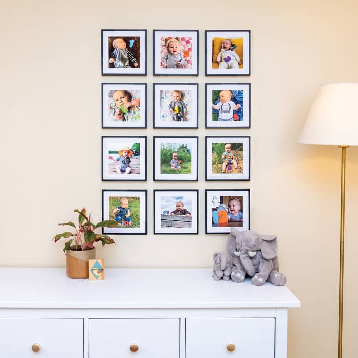 Twelve monthly photos of a baby are displayed in black frames hung in a photo grid above a dresser in a nursery.