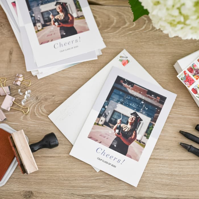 Stamps and pens surround Photo Greeting Cards showing a graduate popping a bottle of champagne.
