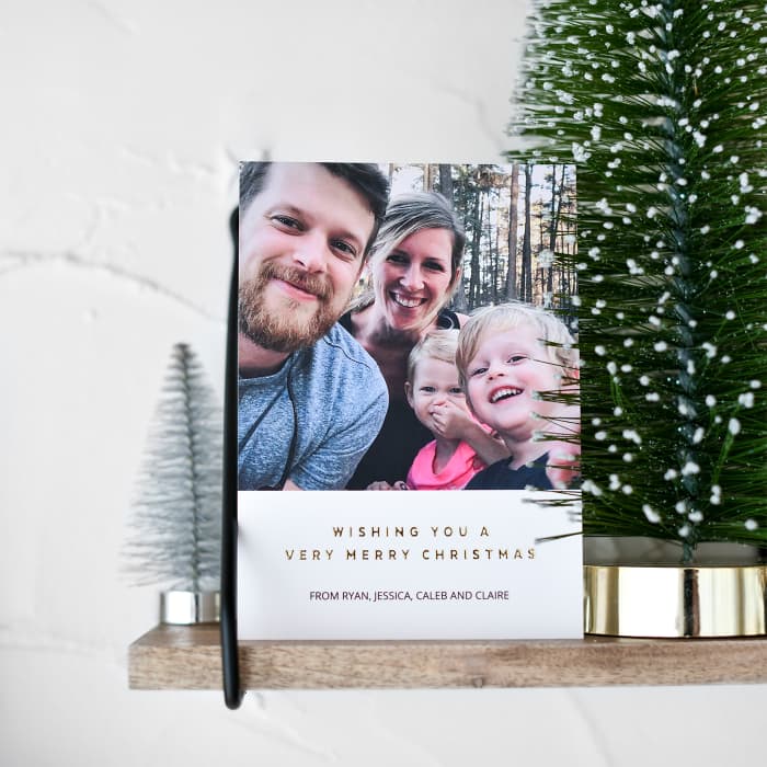 A Holiday Card is propped up on a wooden shelf next to several miniature Christmas trees.