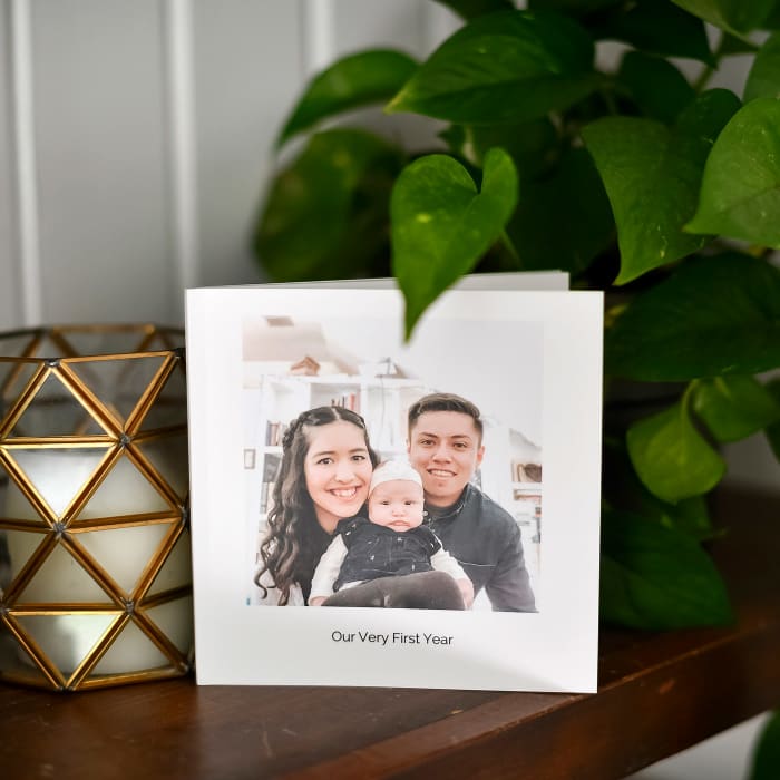 A 6x6 Softcover Photo Book is propped up on a mantle beside a plant and candle. The title reads, Our Baby's First Year.