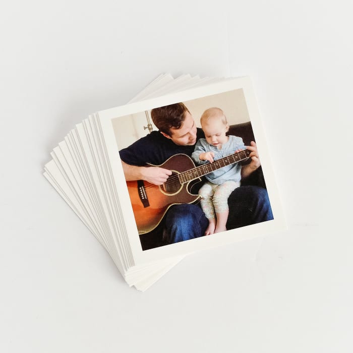 A stack of white bordered Tiny Prints on a white background. The top print shows a toddler learning how to play guitar.