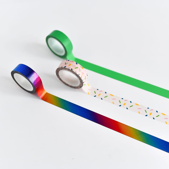 Three rolls of Washi tape are rolled out onto a white desk - bright green, rainbow sprinkle, and metallic rainbow.